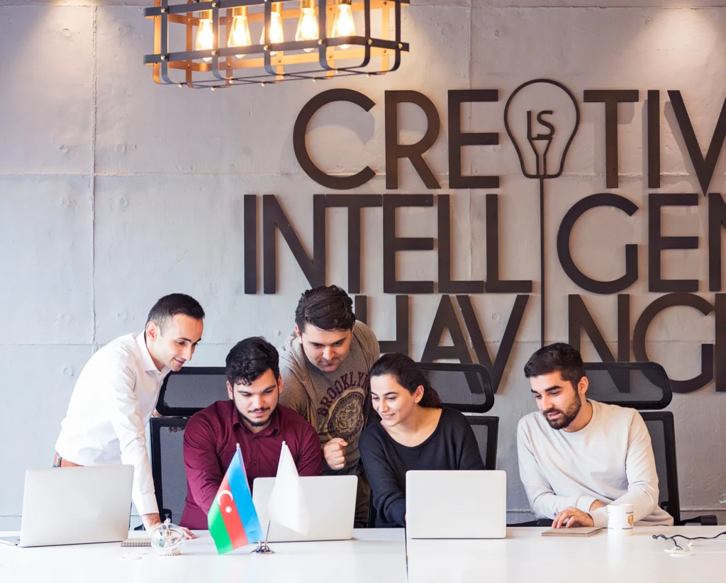 A diverse team of digital marketing professionals collaborating on a laptop in a modern office with "Creative Intelligence" wall art and the Azerbaijan flag.