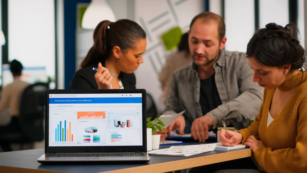 Three people work together at a table with papers and laptops; a nearby laptop screen displays various financial charts and graphs.